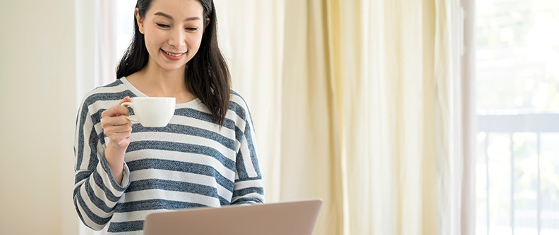 A woman smiles holding a cup and looking at her laptop