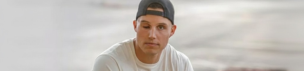 Person sitting in a parking lot wearing black baseball cap backward and white T-shirt. 