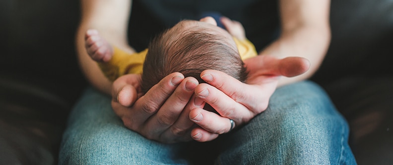 Newborn baby boy with dark brown hair lays cradled in his fathers hands.