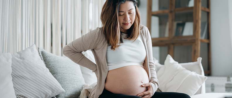 Pregnant woman with her eyes closed touching her belly and lower back, suffering from backache.