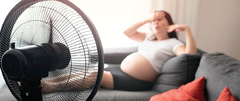 Pregnant person sitting on couch near fan to stay cool.