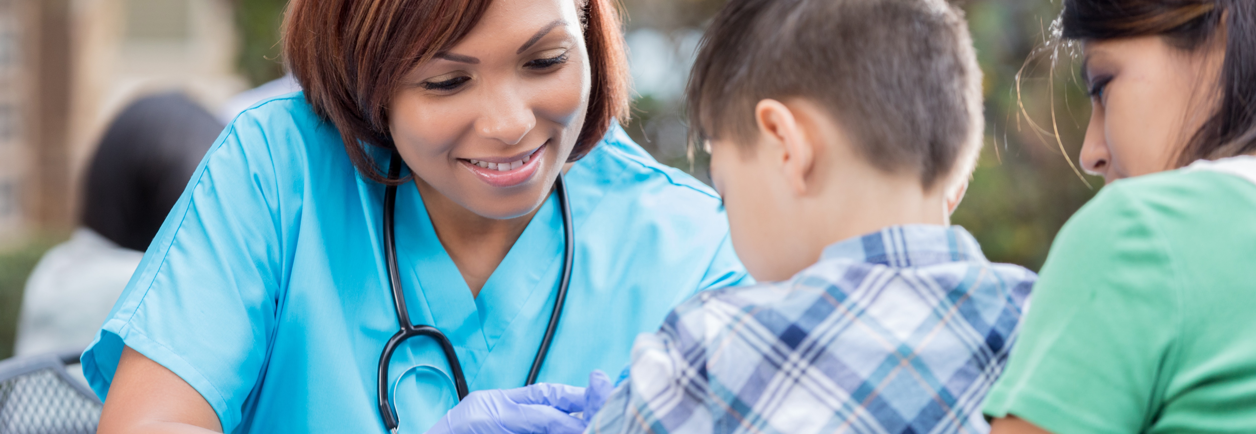 Mid adult African American nurse gives a young boy an immunization at an outdoor free clinic. The boy is sitting in his mom's lap. The nurse is wearing scrubs and a stethoscope.