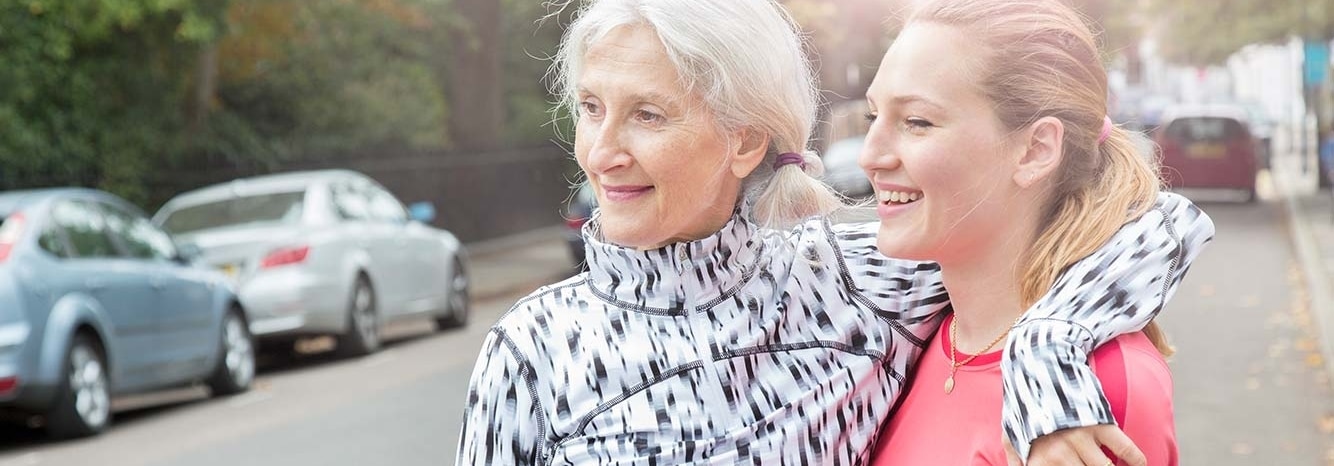 Two women smiling and wearing athletic clothing embrace sideways. One is an older woman with white hair and the other is a younger woman with blond hair.