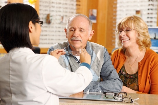 An optometrist holds up glasses for a senior man and his wife, in an optometrist's office