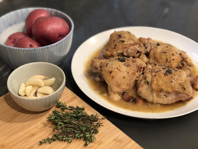 Chicken sitting in broth next to a bowl of garlic cloves and a bowl of potatoes
