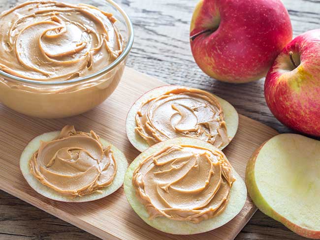 Apple slices topped with nut butter on a cutting board with a bowl of nut butter and whole apples on the side. 