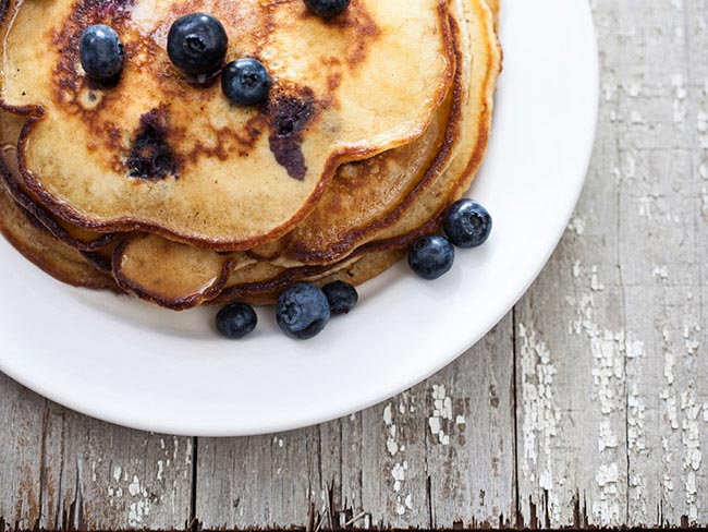 A stack of oatmeal banana pecan blueberry pancakes on a white plate, topped with fresh blueberries.