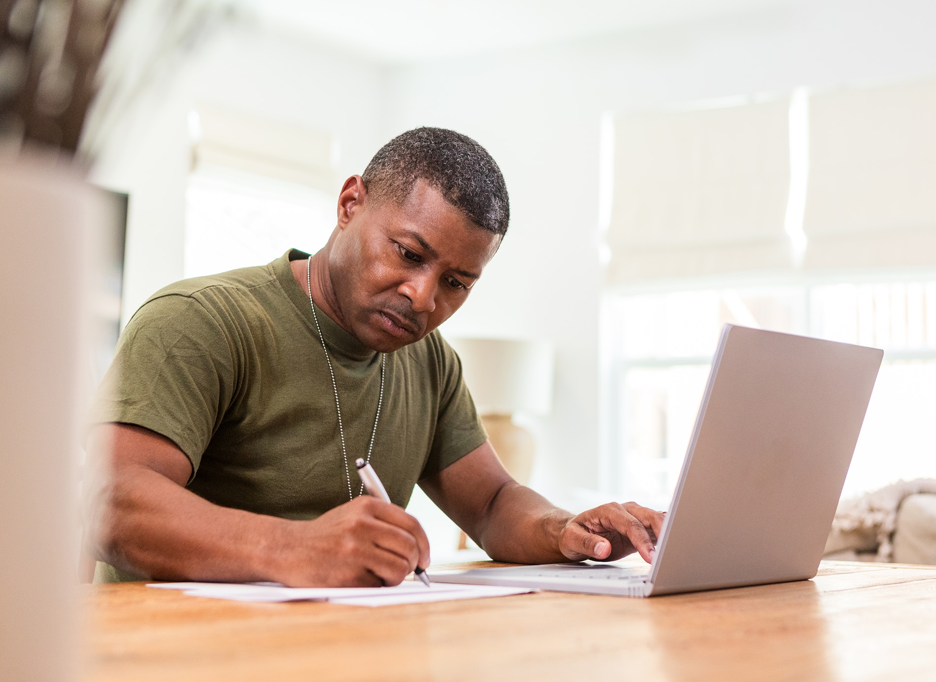 A person sitting at a wooden table, writing notes while using a laptop