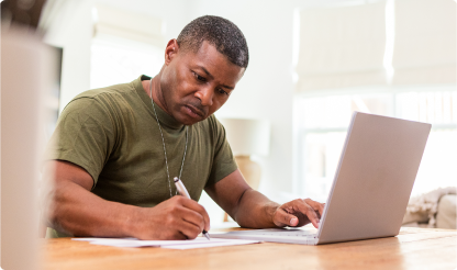 A person sitting at a wooden table, writing notes while using a laptop