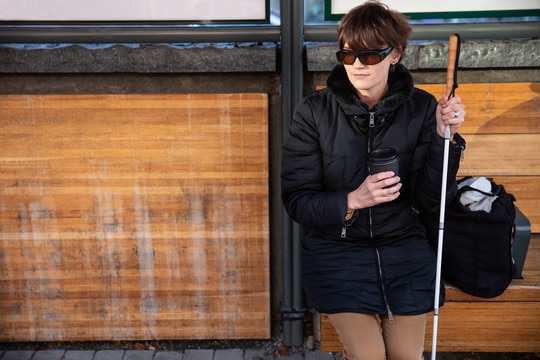Woman holding a white cane in one hand and a coffee cup in the other hand sitting on a bus shelter bench