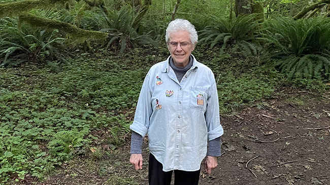 Senior woman with white hair is smiling against a forest backdrop of trees and ferns.