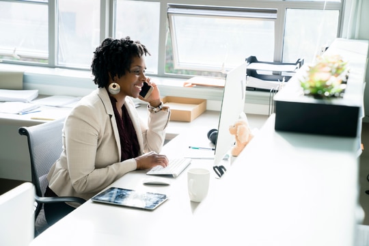 Businesswoman talking on smart phone while using desktop computer in office